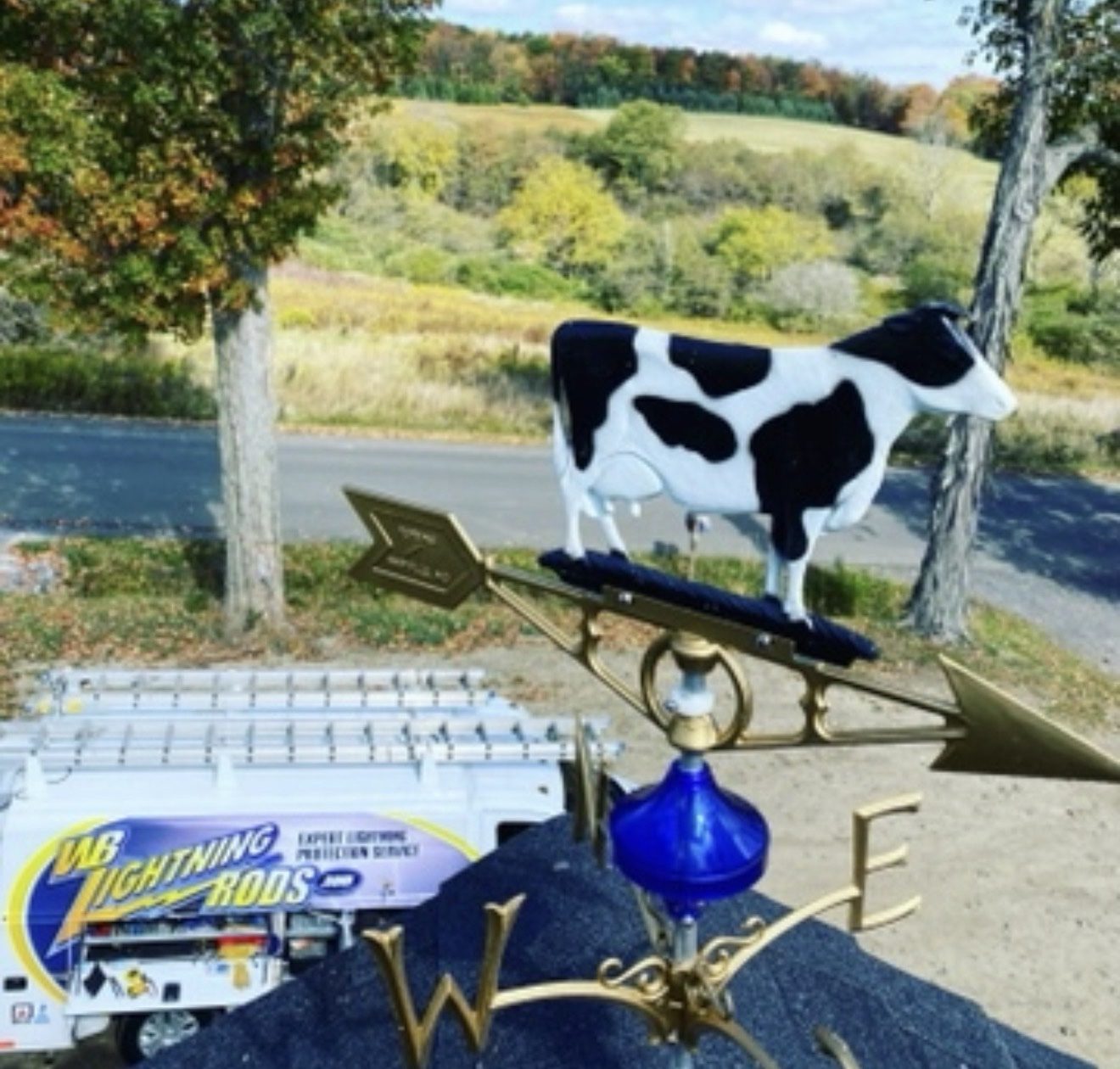 Cow weathervane and glass ball on a dairy barn roof with the WB Lightning Rods van below and farmland behind